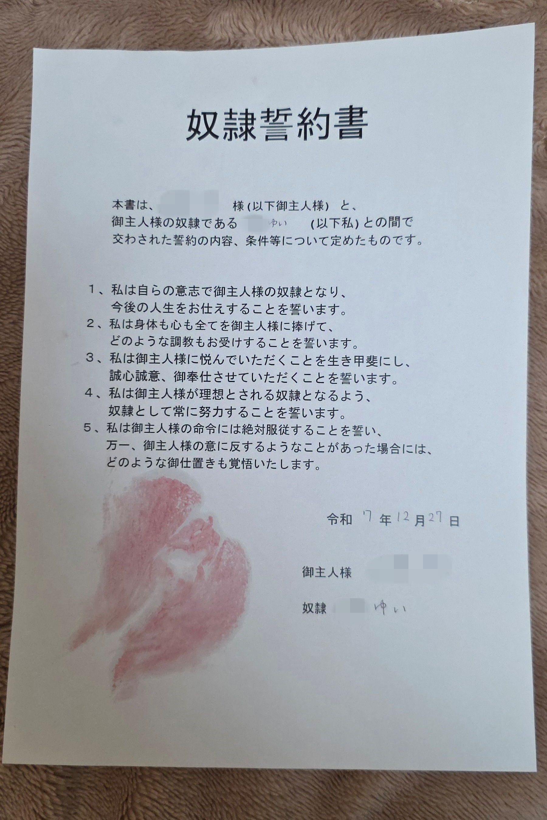 「奴隷誓約書」と題された用紙が置かれている。本文には複数の誓約事項と日付、署名欄があり、用紙の下部には赤い印が残っている。